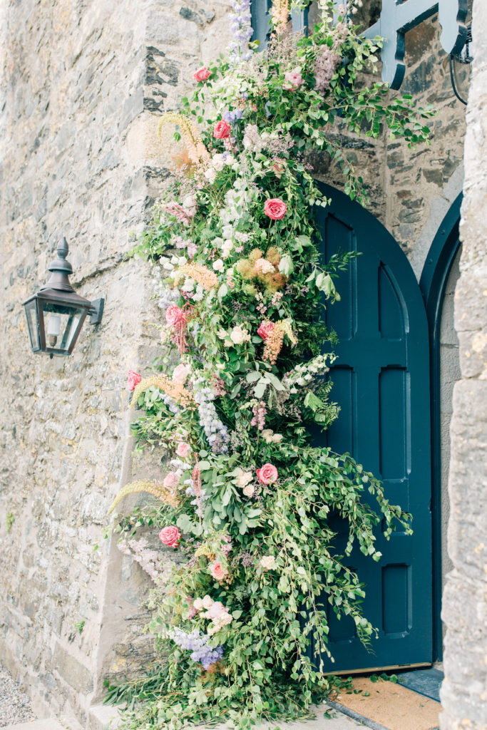 a partial floral arch with pastel flowers over a blue door and stone wall